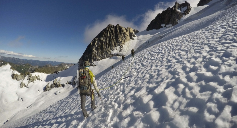 A person wearing a backpack and snow gear, hikes across a snow incline. There are others in front of them. They appear to be at high elevation.